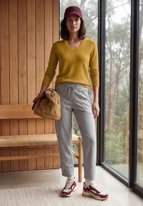 Femme en pull jaune, pantalon gris, chapeau bordeaux, baskets assorties et sac, debout près d’une fenêtre en intérieur boisé.