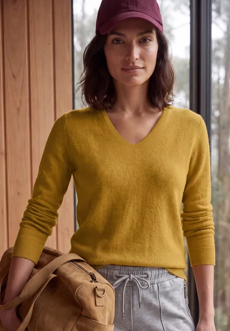 Une femme en pull jaune, pantalon gris et casquette bordeaux tient un sac beige à l'intérieur.