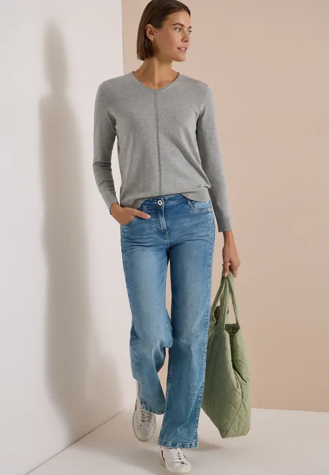 Basic Pullover mit V-Neck in Unifarbe mineral grey melange Frau in grauem V-Pullover, lässigen blauen Jeans, weißen Sneakern und grünem Shopper vor hellem Hintergrund.