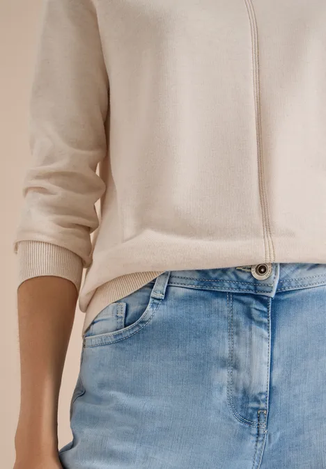 Beige vest en blauwe jeans gedragen door een staand persoon, focus op de taille en het bovenlichaam.