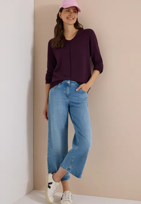 Een jonge vrouw draagt een paarse blouse, wijde blauwe jeans, witte sneakers en een roze pet.