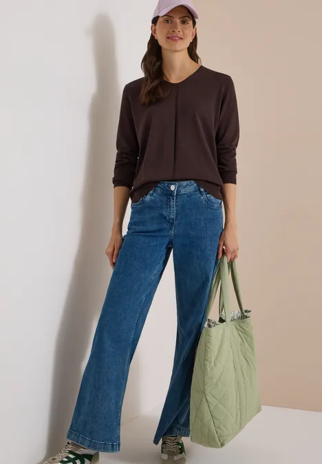 Eenvoudige trui met V-hals in effen kleur truffle brown Vrouw in donkerbruin shirt, wijde jeans met split vooraan, witte sneakers en een grote groene tas.