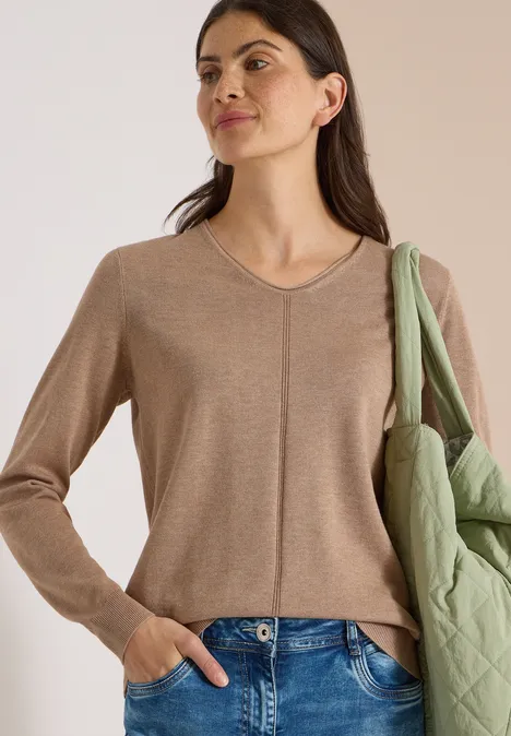 Eenvoudige trui met V-hals in effen kleur creamy mocha melange Vrouw in beige V-trui en blauwe jeans, met een groen gewatteerd jack losjes over de schouder.