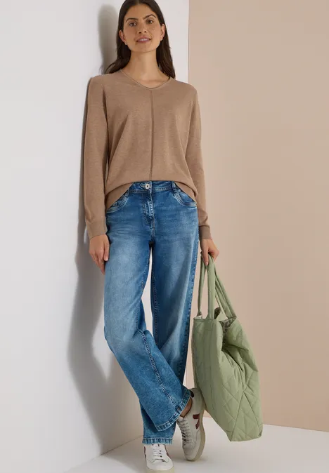 Eenvoudige trui met V-hals in effen kleur creamy mocha melange Vrouw in beige trui, blauwe jeans, witte sneakers met grote groene gewatteerde tas tegen neutrale achtergrond.