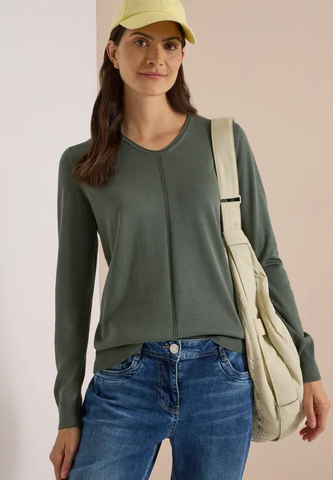 Eenvoudige trui met V-hals in effen kleur dried olive green Een jonge vrouw in een groen shirt met lange mouwen, spijkerbroek, gele pet en beige schoudertas.