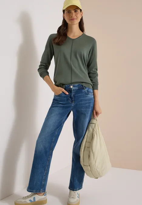 Eenvoudige trui met V-hals in effen kleur dried olive green Vrouw in groene trui, blauwe jeans, witte sneakers en gele pet, met een lichte tas in de hand.