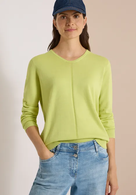 Eenvoudige trui met V-hals in effen kleur citrin green Vrouw in lichtgroene trui, spijkerbroek en pet voor een neutrale achtergrond.