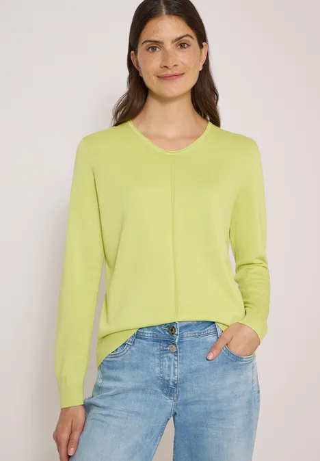 Eenvoudige trui met V-hals in effen kleur citrin green Vrouw met lichtgroene trui en blauwe jeans, hand in de zak, glimlachend voor een effen achtergrond.