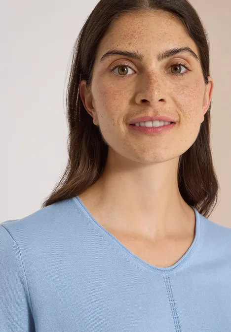 Eenvoudige trui met V-hals in effen kleur easy blue Portret van een glimlachende vrouw met bruin haar in een lichtblauw shirt tegen neutrale achtergrond.