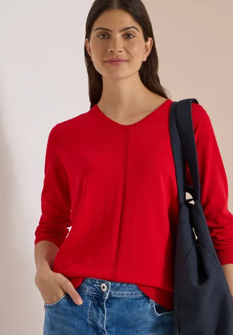 Eenvoudige trui met V-hals in effen kleur flame red Vrouw met bruin haar draagt een rode trui en donkerblauwe tas over haar schouder voor een lichte achtergrond.