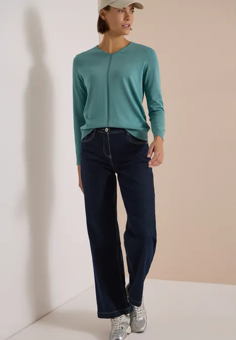 Basic Pullover mit V-Neck in Unifarbe mineral blue Eine Frau in grüner Strickjacke, Jeans und Sneakern vor hellem Hintergrund.