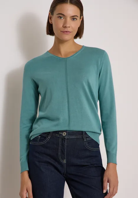 Basic Pullover mit V-Neck in Unifarbe mineral blue Frau trägt tealfarbenen Pullover und dunkle Jeans, steht vor hellem Hintergrund.