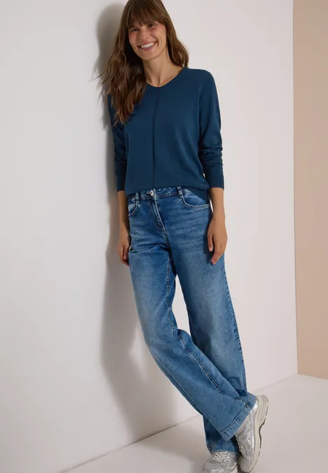Basic Pullover mit V-Neck in Unifarbe moonlight blue Eine lächelnde Frau in Jeans, Sneakern und blauem Oberteil lehnt an einer Wand.