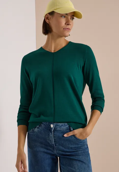 Pull couleur unie à col en V dark fir green Femme en casquette jaune, pull vert et jean bleu, posant de profil devant un fond clair.