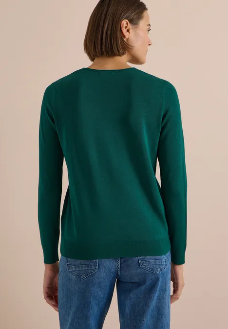 Pull couleur unie à col en V dark fir green Vue de dos d’une femme portant un pull vert et un jean bleu sur fond beige.