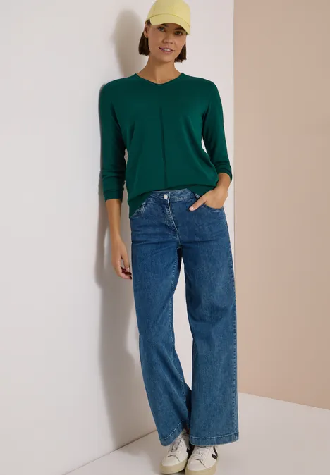 Pull couleur unie à col en V dark fir green Une femme posant contre un mur, vêtue d’un gilet vert, d’un jean large et d’une casquette jaune.