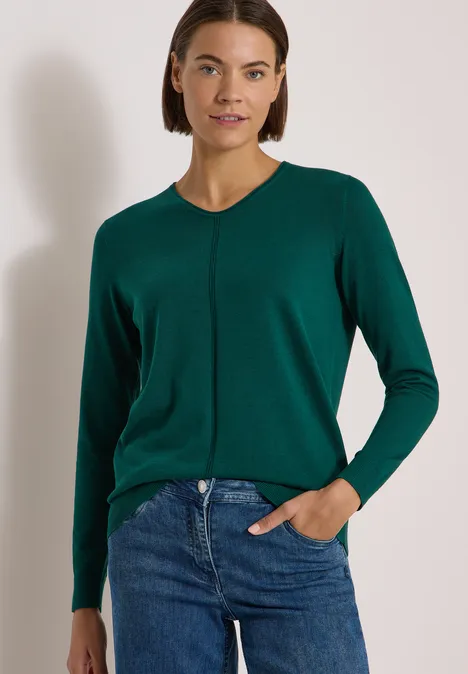 Pull couleur unie à col en V dark fir green Femme en pull vert et jean, posant de manière décontractée sur fond neutre.