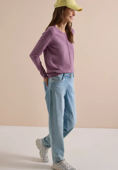 Eine Frau mit grüner Cap, lila Pullover, hellblauen Jeans und Sneakern lächelt entspannt mit Händen in den Taschen.