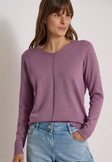 Junge Frau im violetten Pullover mit vertikalem Ziernaht und Jeans, Hände in Hosentaschen.