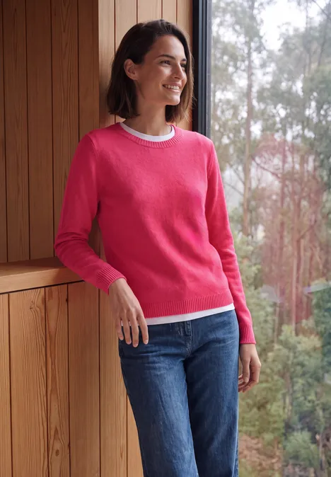 Effen trui met boothals cosy coral melange Vrouw in roze trui en jeans lacht voor een houten kader met uitzicht op het bos.