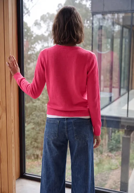 Effen trui met boothals cosy coral melange Achteraanzicht van persoon in roze trui en blauwe jeans voor een raam met uitzicht op bos.
