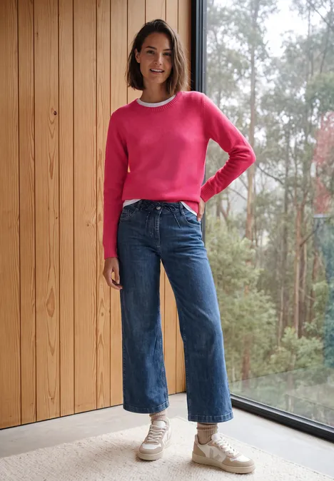 Effen trui met boothals cosy coral melange Vrouw in roze trui en wijde jeans voor houten wand en raam met uitzicht op bos.