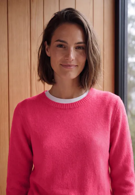 Effen trui met boothals cosy coral melange Vrouw met bruin haar lacht en draagt een roze trui voor een houten wand.