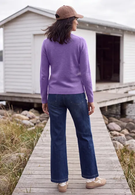 Vrouw in paarse trui, spijkerbroek en pet staat op een houten loopbrug voor een wit huisje.