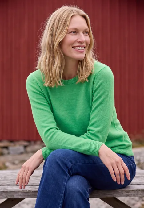 Pullover mit Boat Neck in Unifarbe celery green melange Eine lächelnde Frau in grünem Pullover und Jeans sitzt entspannt auf einer Bank vor roter Hauswand.