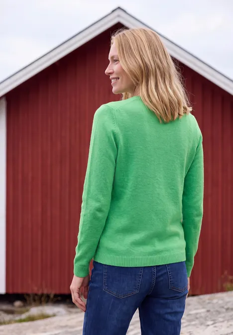 Pullover mit Boat Neck in Unifarbe celery green melange Frau in grünem Pullover und Jeans steht mit dem Rücken zur Kamera vor einer roten Holzwand.