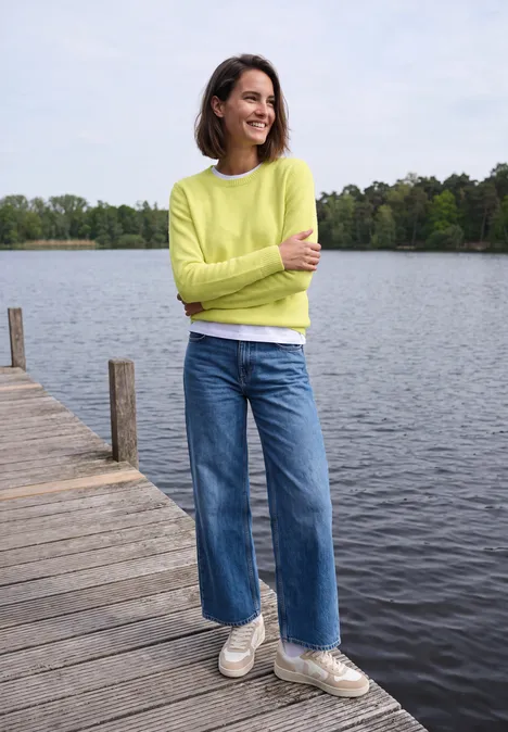 Pullover mit Boat Neck in Unifarbe iced yellow melange Frau steht auf einem Steg am See, trägt einen hellgrünen Pullover, Jeans und weiße Sneaker, lächelt.