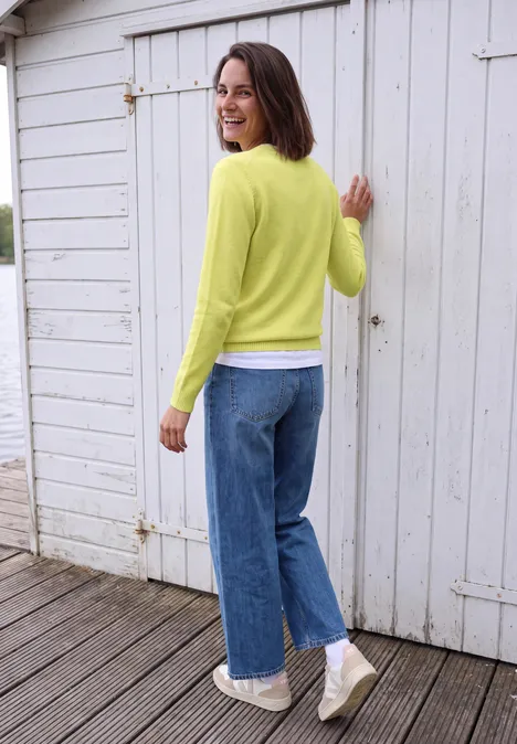 Pullover mit Boat Neck in Unifarbe iced yellow melange Frau im gelben Pullover und Jeans steht lächelnd auf Holzsteg vor weißem Häuschen.