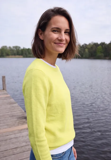 Pullover mit Boat Neck in Unifarbe iced yellow melange Eine lächelnde Frau in gelbem Pullover steht auf einem Steg am See mit Bäumen im Hintergrund.
