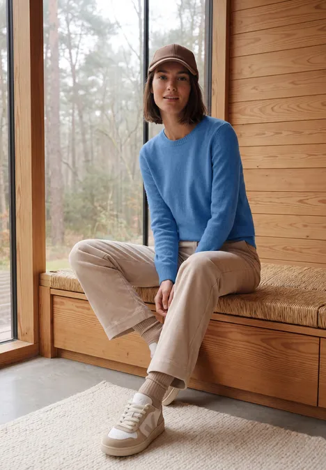 Effen trui met boothals north blue melange Vrouw in blauwe trui en pet zit op een houten bank in een lichte ruimte met uitzicht op het bos.