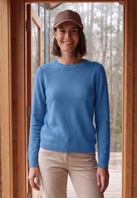 Effen trui met boothals north blue melange Lachende vrouw in blauw truitje, beige broek en pet, staand voor een houten raam.