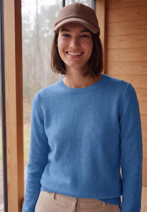 Effen trui met boothals north blue melange Jonge vrouw met blauwe trui en beige pet voor een houten wand, ze lacht.
