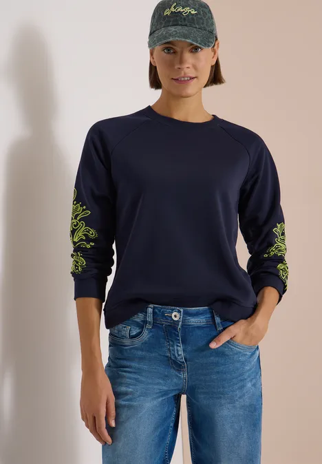 Sweatshirt mit Artwork am Ärmel urban dark blue Frau in navyblauem Sweatshirt mit neongrünen Blumenstickereien an den Ärmeln, Jeans und Basecap.