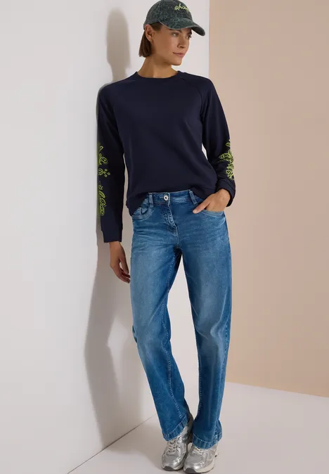 Sweatshirt mit Artwork am Ärmel urban dark blue Frau in dunkelblauem Pullover mit grünen Ärmeldetails, blauen Jeans, Cap und Sneakern.
