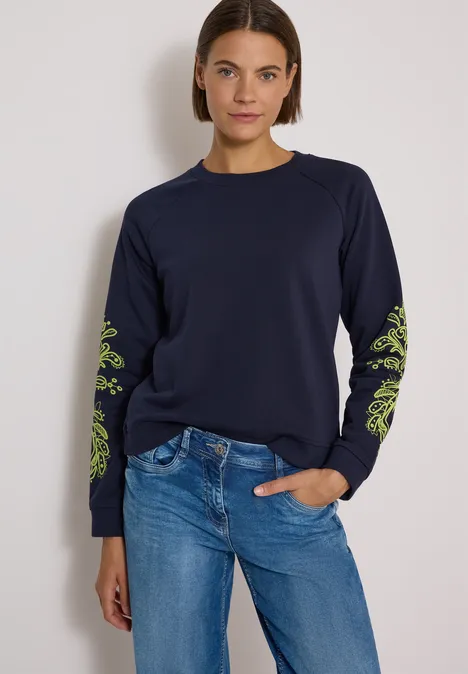 Sweatshirt mit Artwork am Ärmel urban dark blue Frau in dunkelblauem Sweatshirt mit grünen Ärmelmustern und Jeans vor hellem Hintergrund.