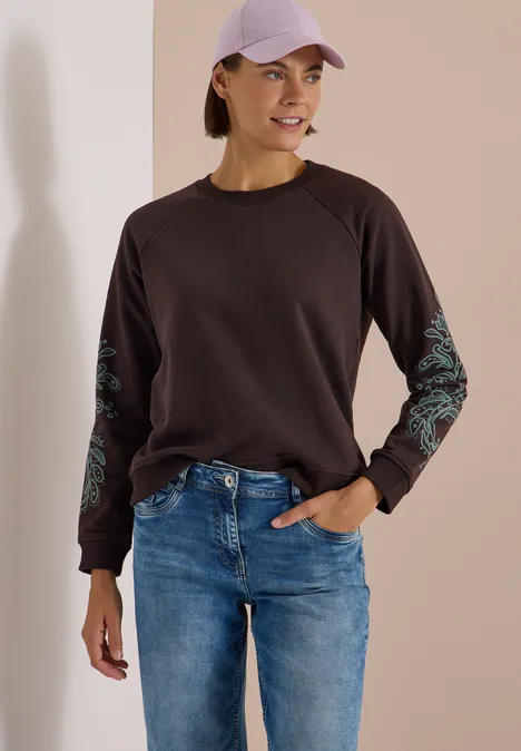 Sweatshirt mit Artwork am Ärmel truffle brown Frau in lässigem Outfit: dunkles Sweatshirt mit Musterärmeln, Jeans und lila Cap, lächelnd vor neutralem Hintergrund.