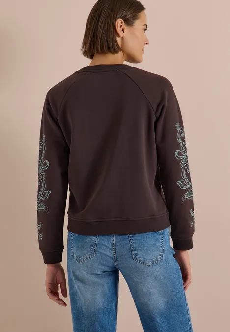 Sweatshirt mit Artwork am Ärmel truffle brown Frau von hinten in dunkelbraunem Sweatshirt mit türkisfarbener Blumenstickerei an Ärmeln und blauen Jeans.