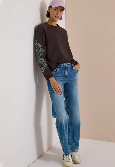 Sweatshirt mit Artwork am Ärmel truffle brown Frau lehnt an Wand, trägt schwarzes Sweatshirt mit türkis Muster auf dem Ärmel, blaue Jeans, weiße Sneaker und rosa Cap.