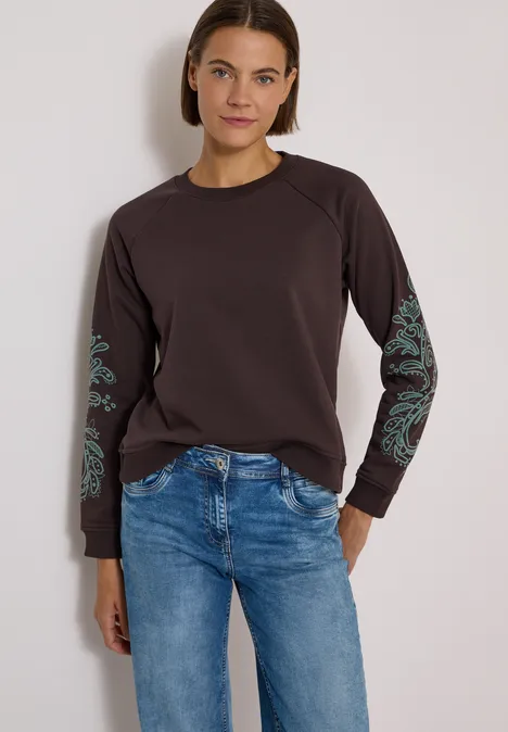 Sweatshirt mit Artwork am Ärmel truffle brown Frau in Jeans und braunem Langarmshirt mit grünen Mustern an den Ärmeln vor neutralem Hintergrund.