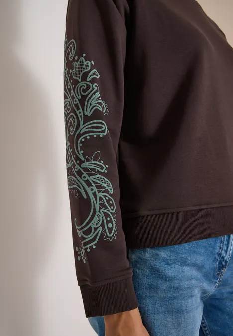 Sweatshirt mit Artwork am Ärmel truffle brown Schwarzer Pullover mit türkisem Paisleymuster am Ärmel und blaue Jeans.