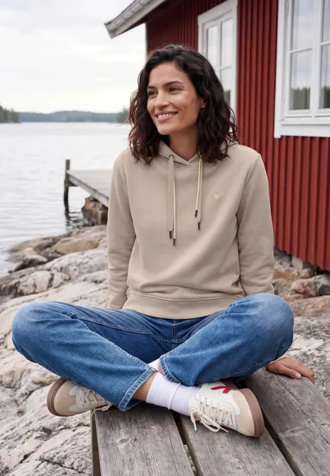 Femme assise en tailleur sur un quai en bois devant une maison rouge au bord d'un lac.