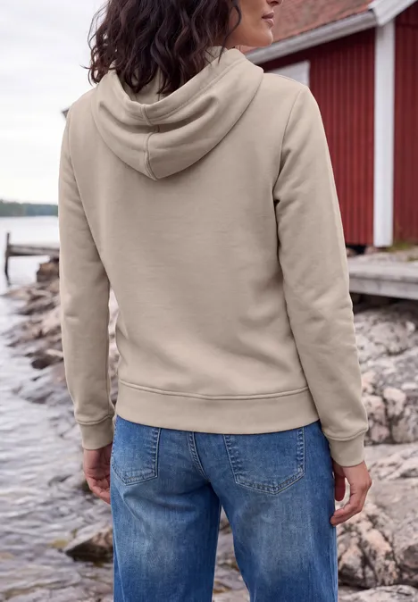 Personne portant un sweat à capuche beige et jean, debout sur un rivage rocheux devant un hangar à bateaux rouge.