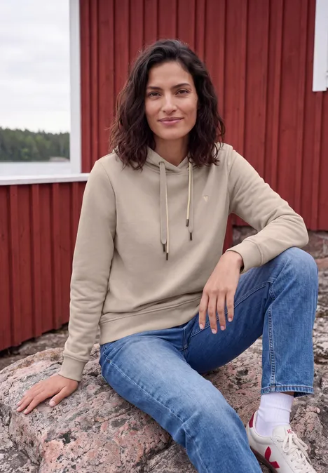 Une femme en sweat à capuche beige et jean est assise sur un rocher devant un mur de bois rouge, souriante.
