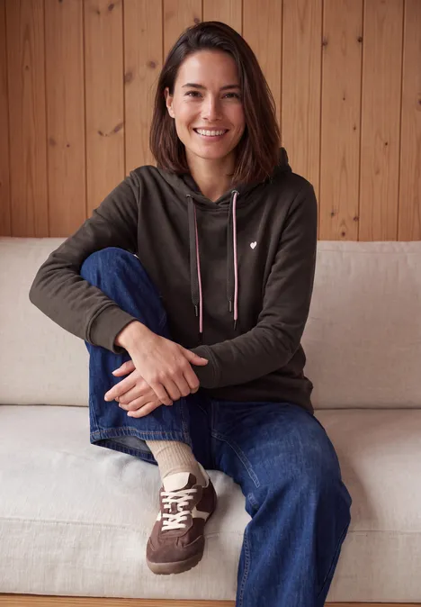 Langarm Sweatshirt mit Kapuze truffle brown Eine Frau sitzt entspannt auf einem hellen Sofa vor einer Holzverkleidung, trägt Hoodie, Jeans und Sneakers.