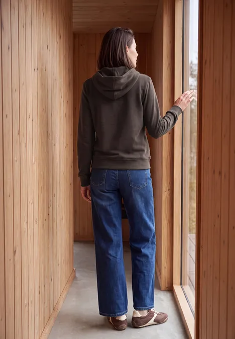 Langarm Sweatshirt mit Kapuze truffle brown Frau in Hoodie und Jeans, von hinten in einem Holzraum stehend, blickt aus einem großen Fenster.