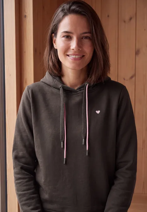 Langarm Sweatshirt mit Kapuze truffle brown Eine lächelnde Frau in dunkelgrauer Kapuzenjacke mit kleinem Herzlogo vor Holzpaneelen.